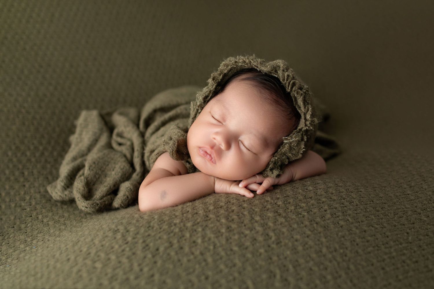 Sleeping newborn baby in olive green knit bonnet and wrap on matching green textured backdrop.