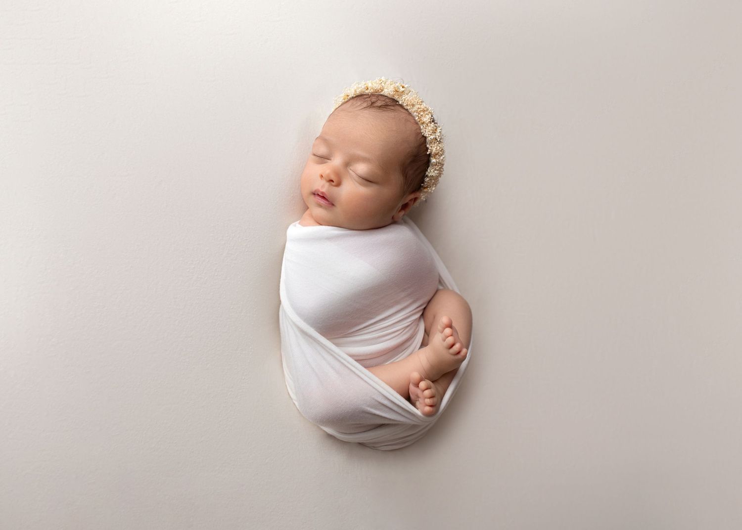 Sleeping newborn baby swaddled in white wrap wearing a delicate floral headband on a white background.