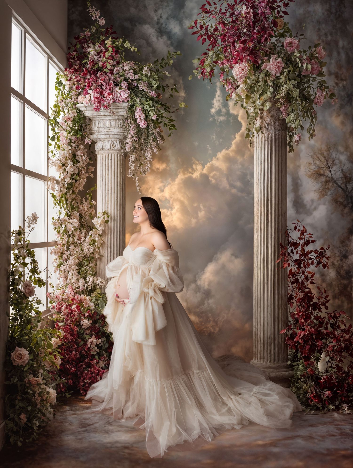 Pregnant woman in cream tulle gown stands between floral columns with dramatic cloudy sky backdrop.