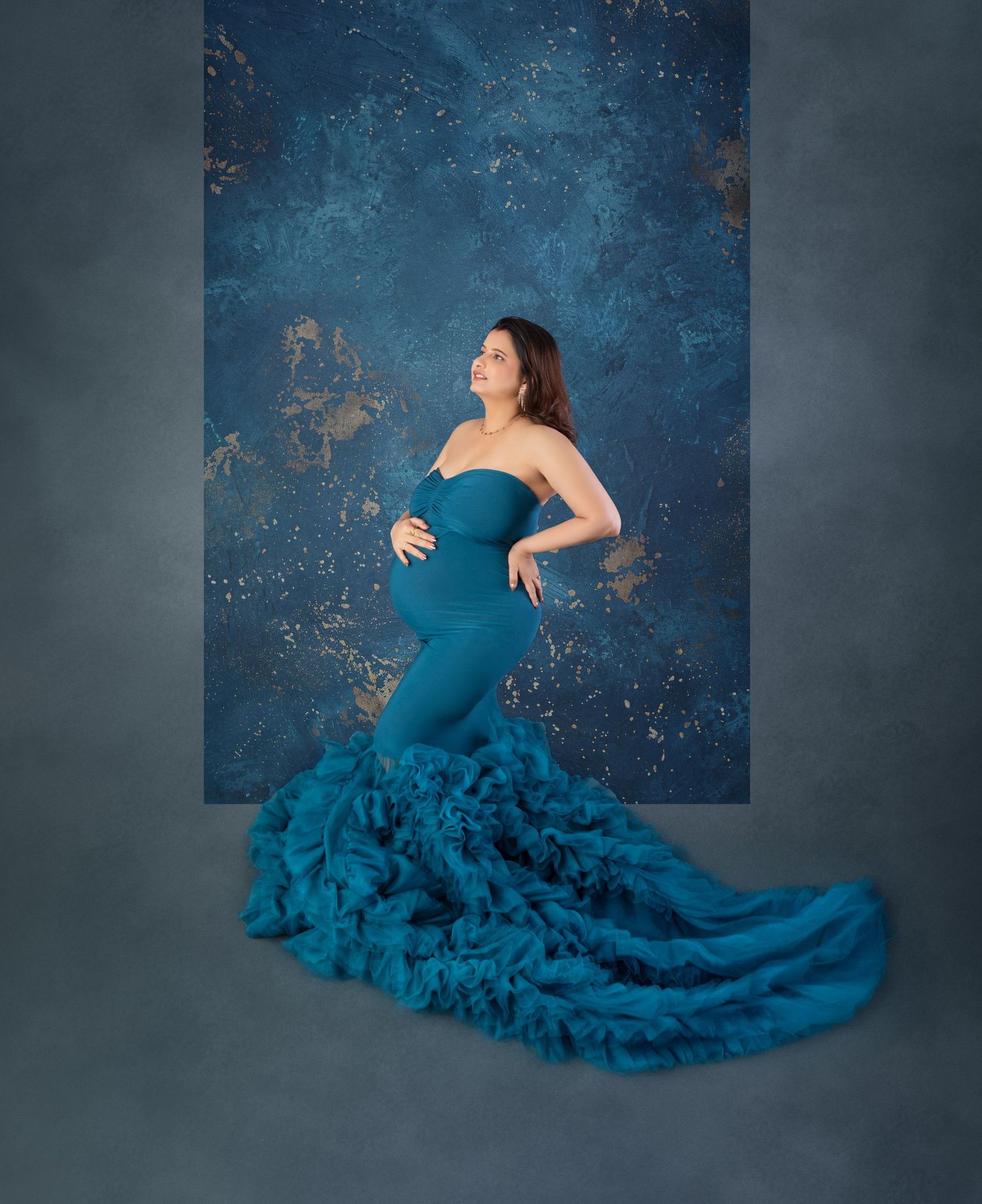 Pregnant woman in strapless teal mermaid gown with ruffle train poses against blue textured backdrop.