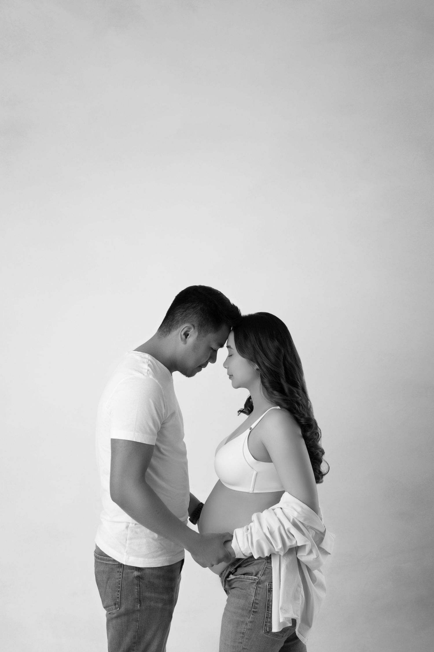 Black and white side profile of pregnant couple touching foreheads tenderly while holding baby bump.