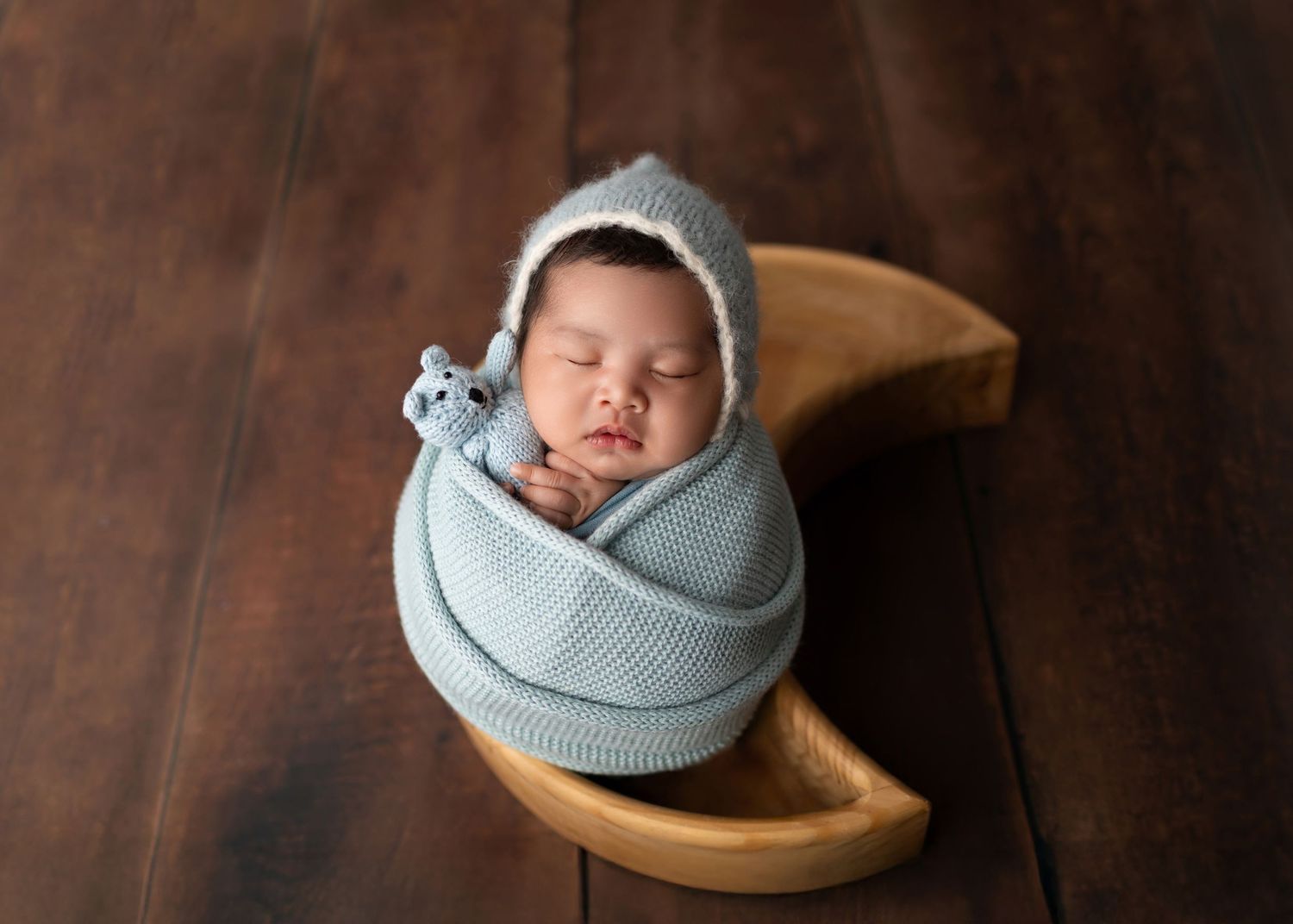 Newborn baby wrapped in blue knit blanket sleeping in a wooden moon prop with tiny bear.
