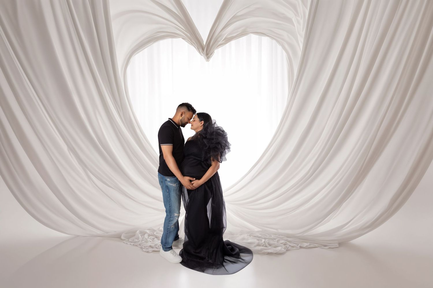 Couple sharing a tender moment in a maternity photo shoot, framed by elegant white draped fabric forming a heart shape.