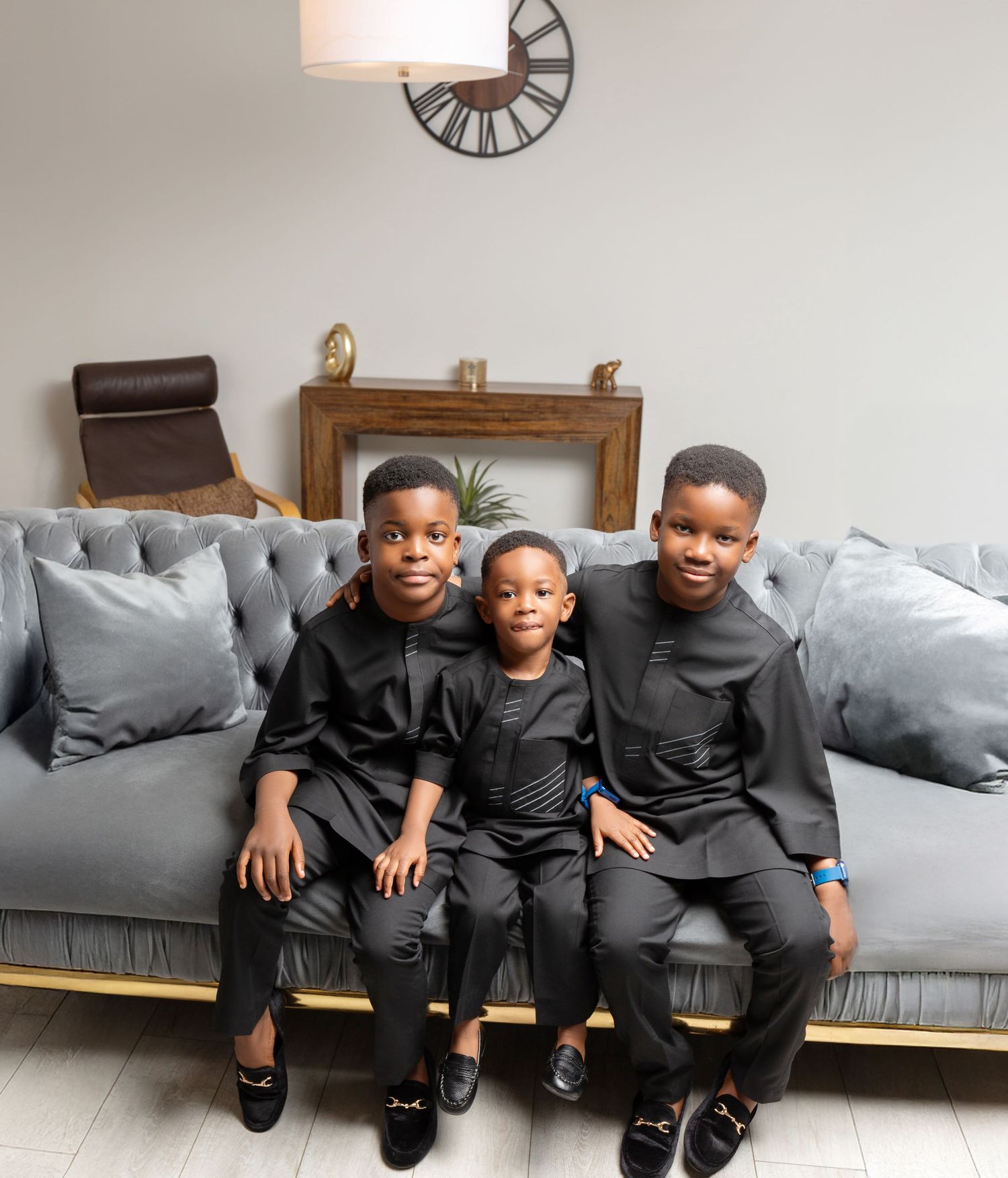 Three young boys in matching black outfits sitting together on a gray tufted sofa in a modern living room.