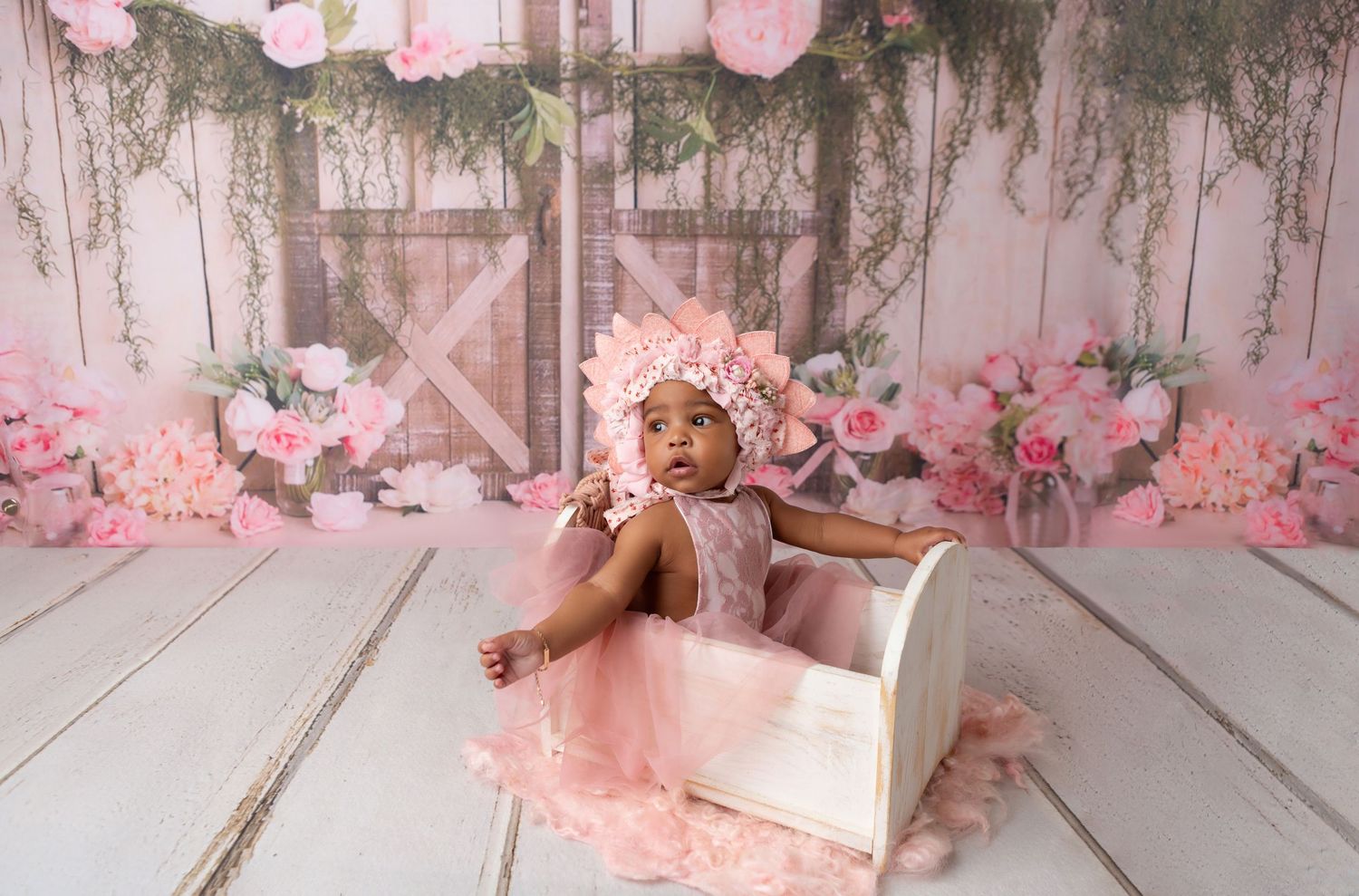 Baby girl in pink dress and floral bonnet sitting in white wooden bed with pink roses backdrop.