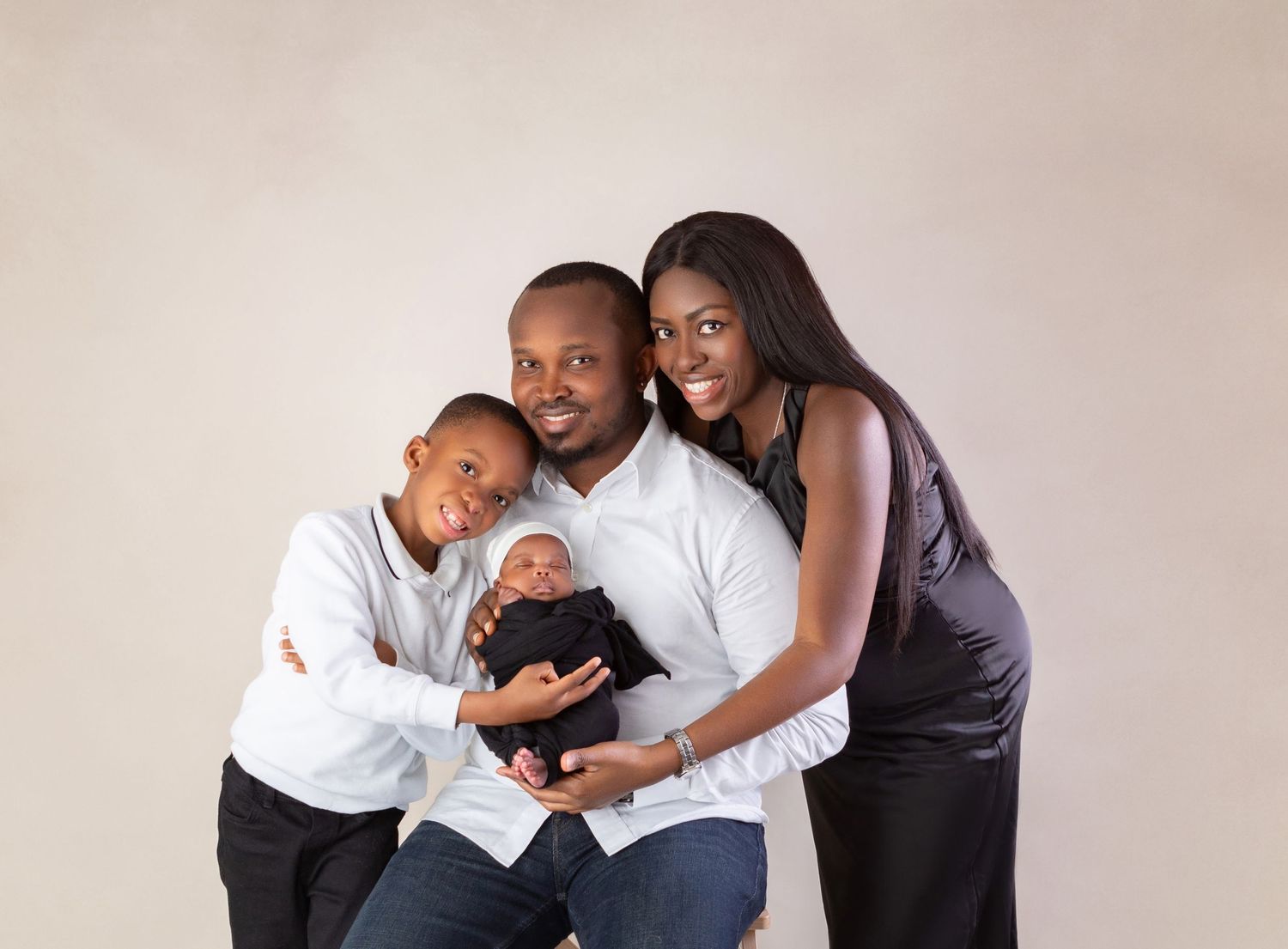 A joyful Black family poses in a studio, parents holding their newborn baby alongside their young son in formal attire.