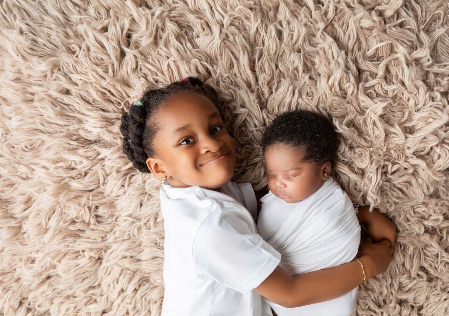 Sweet Black girl lying on fluffy beige rug tenderly holding sleeping newborn baby sibling.