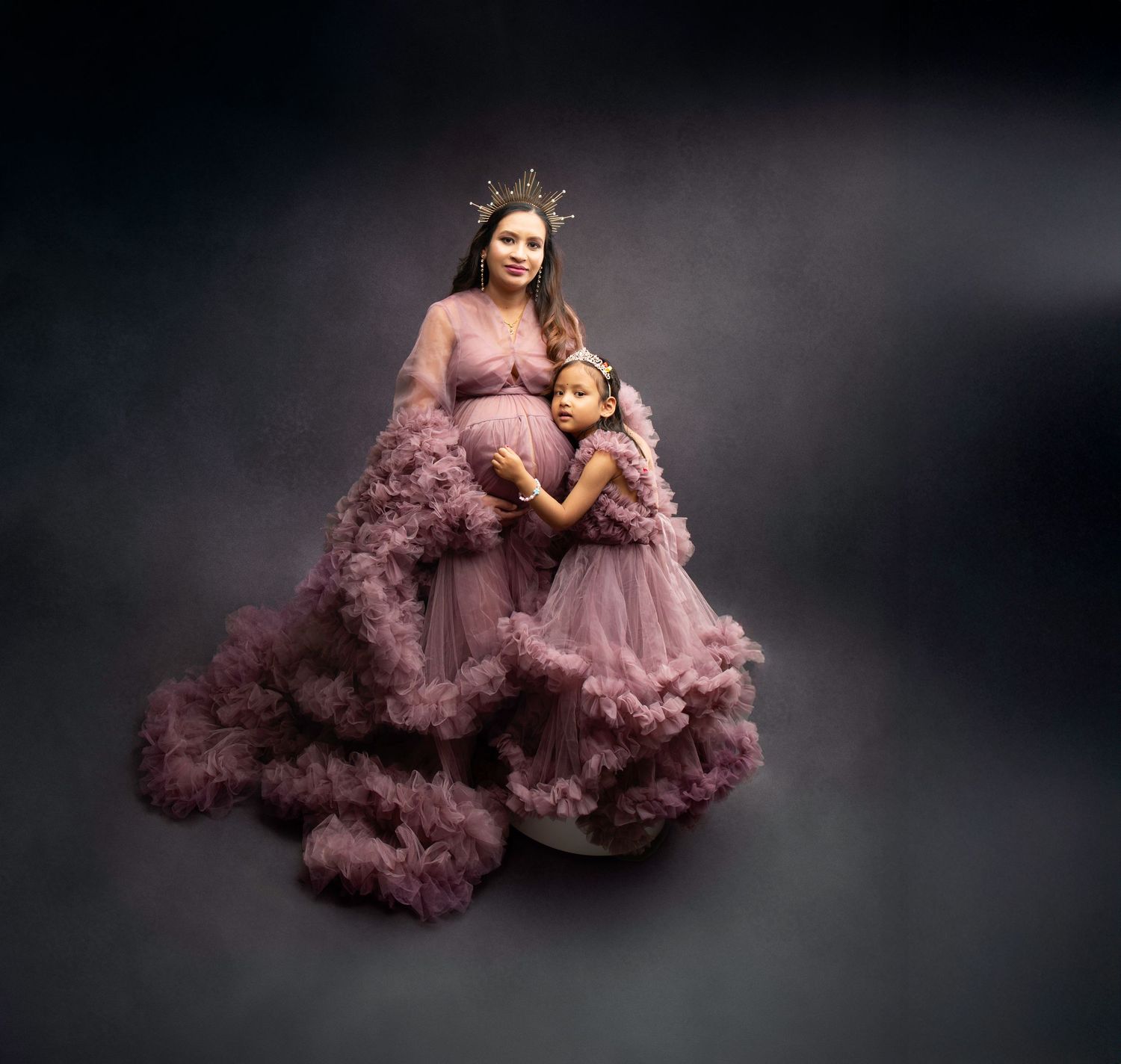 Pregnant woman wearing crown and mauve tulle gown poses with young daughter in matching dress.