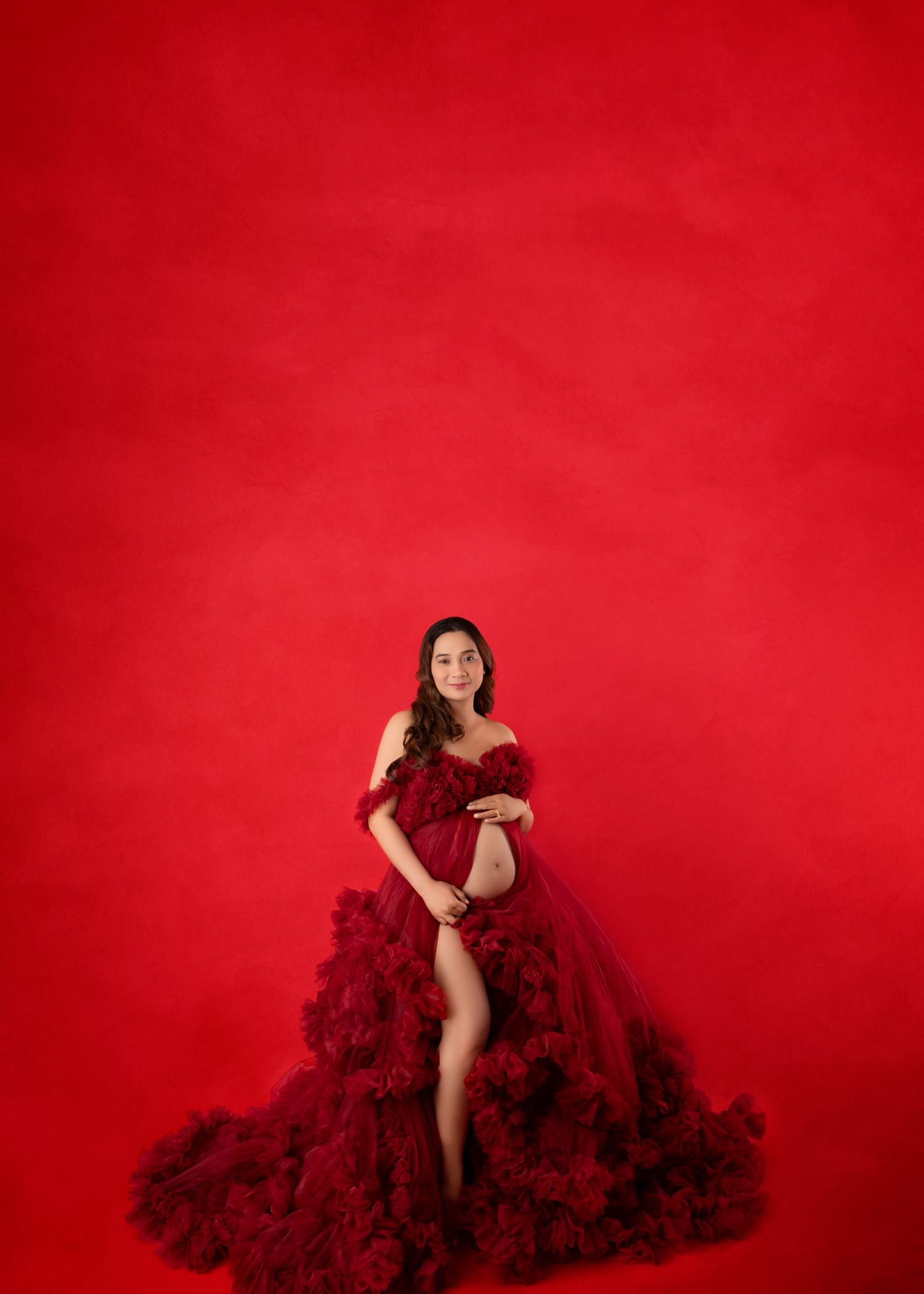 Pregnant woman in elegant red ruffled off-shoulder gown poses gracefully against a bold red studio backdrop.