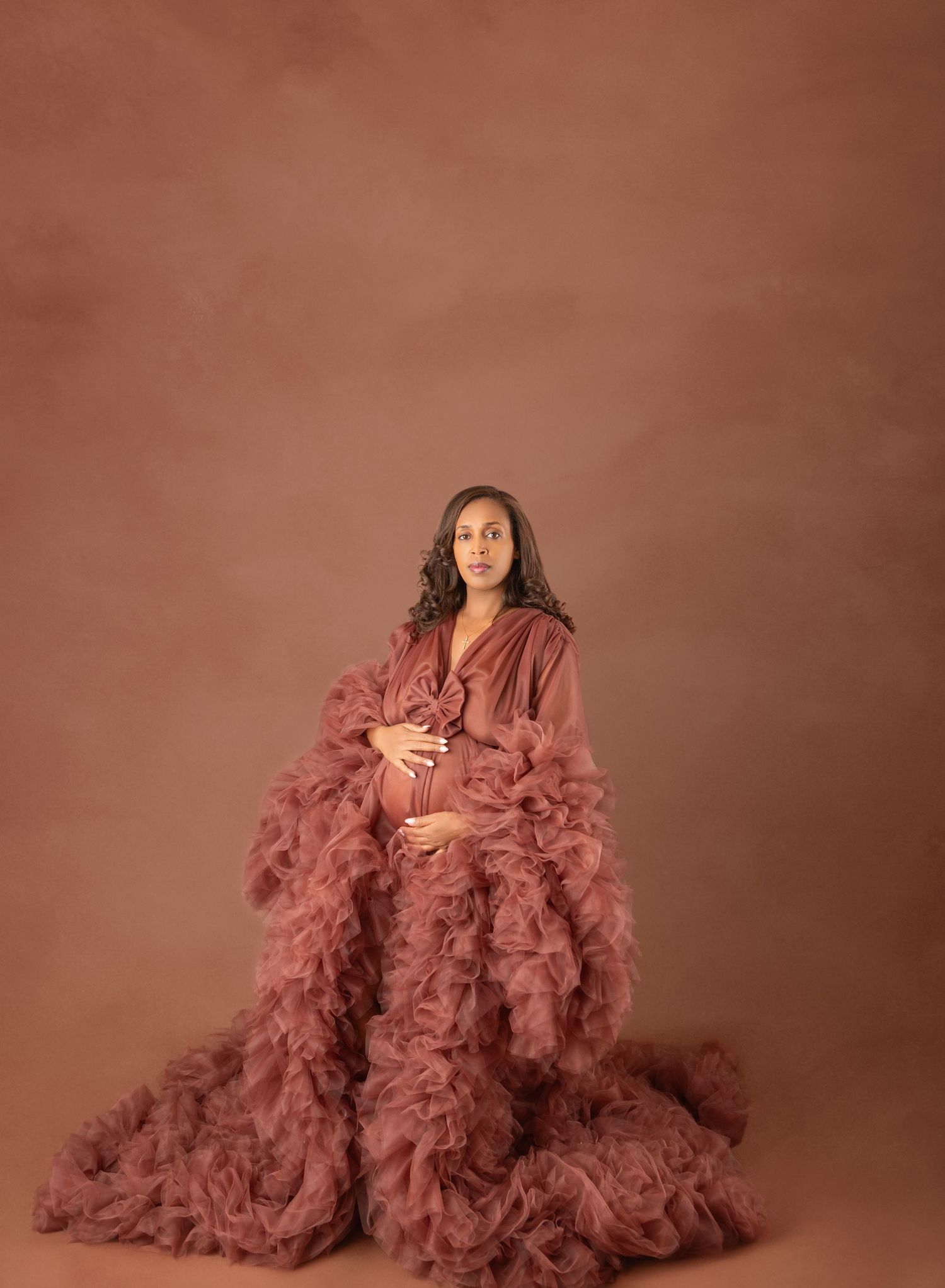 Pregnant woman in voluminous dusty rose ruffled maternity robe in warm-toned studio.