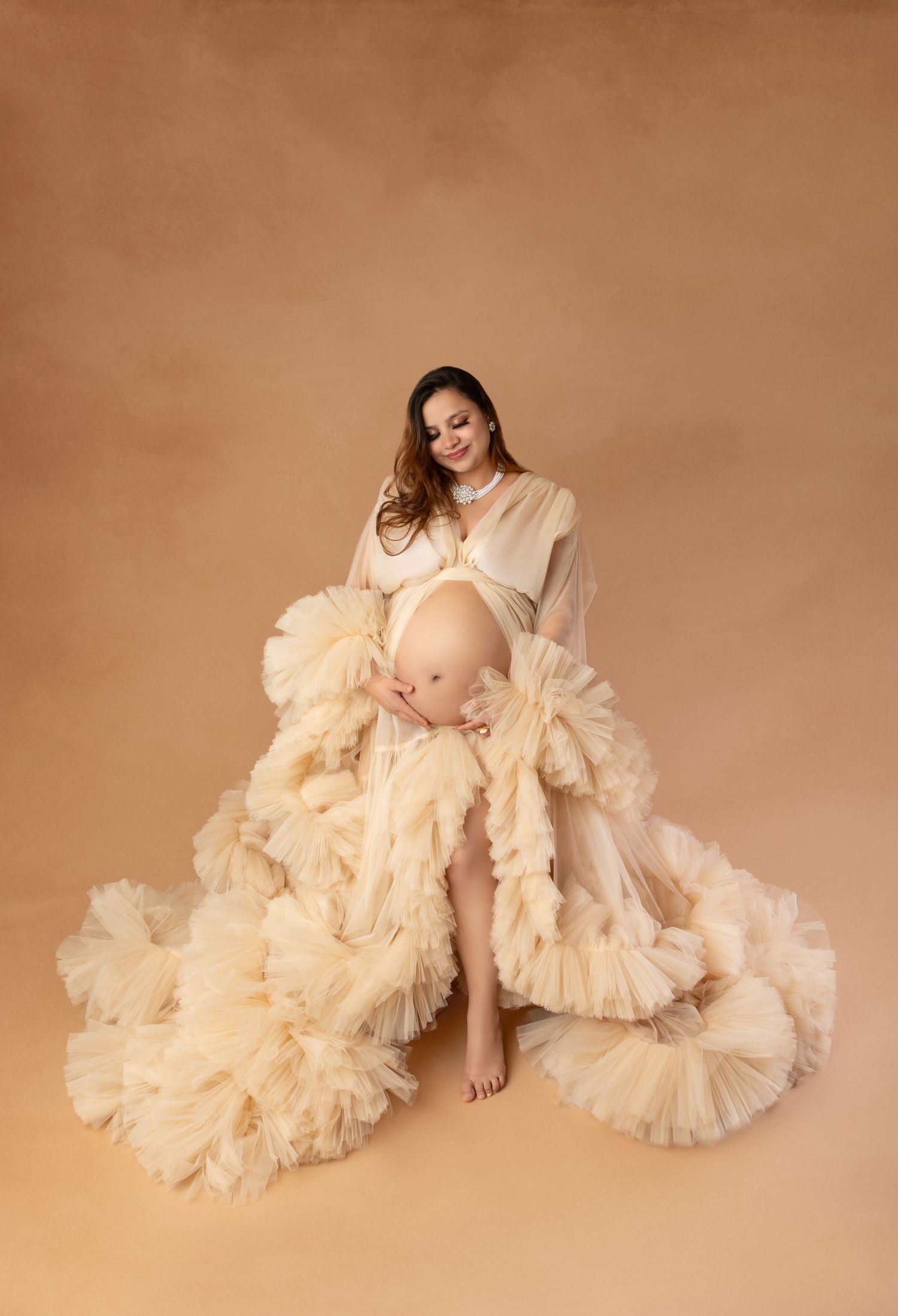 Pregnant woman in flowing cream ruffle tulle maternity gown looking down at baby bump in studio.
