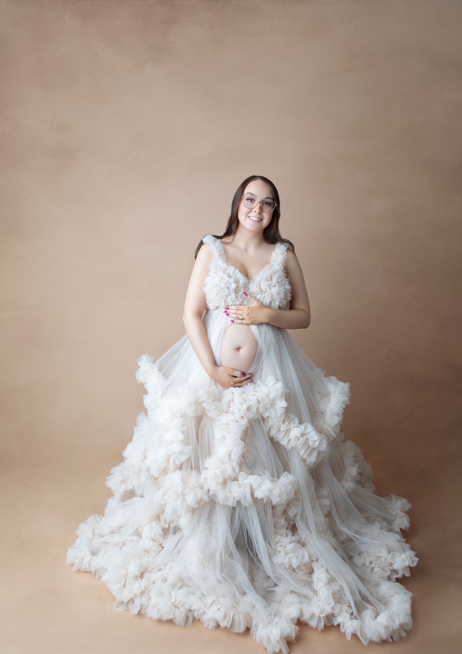 Smiling pregnant woman in white ruffled tulle maternity gown with glasses, studio.