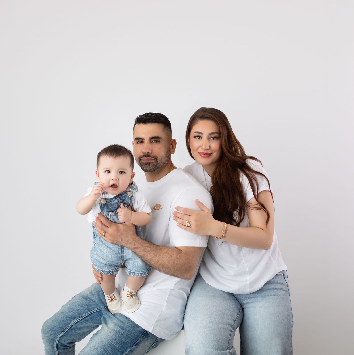 Parents holding smiling baby in denim overalls against white studio background.