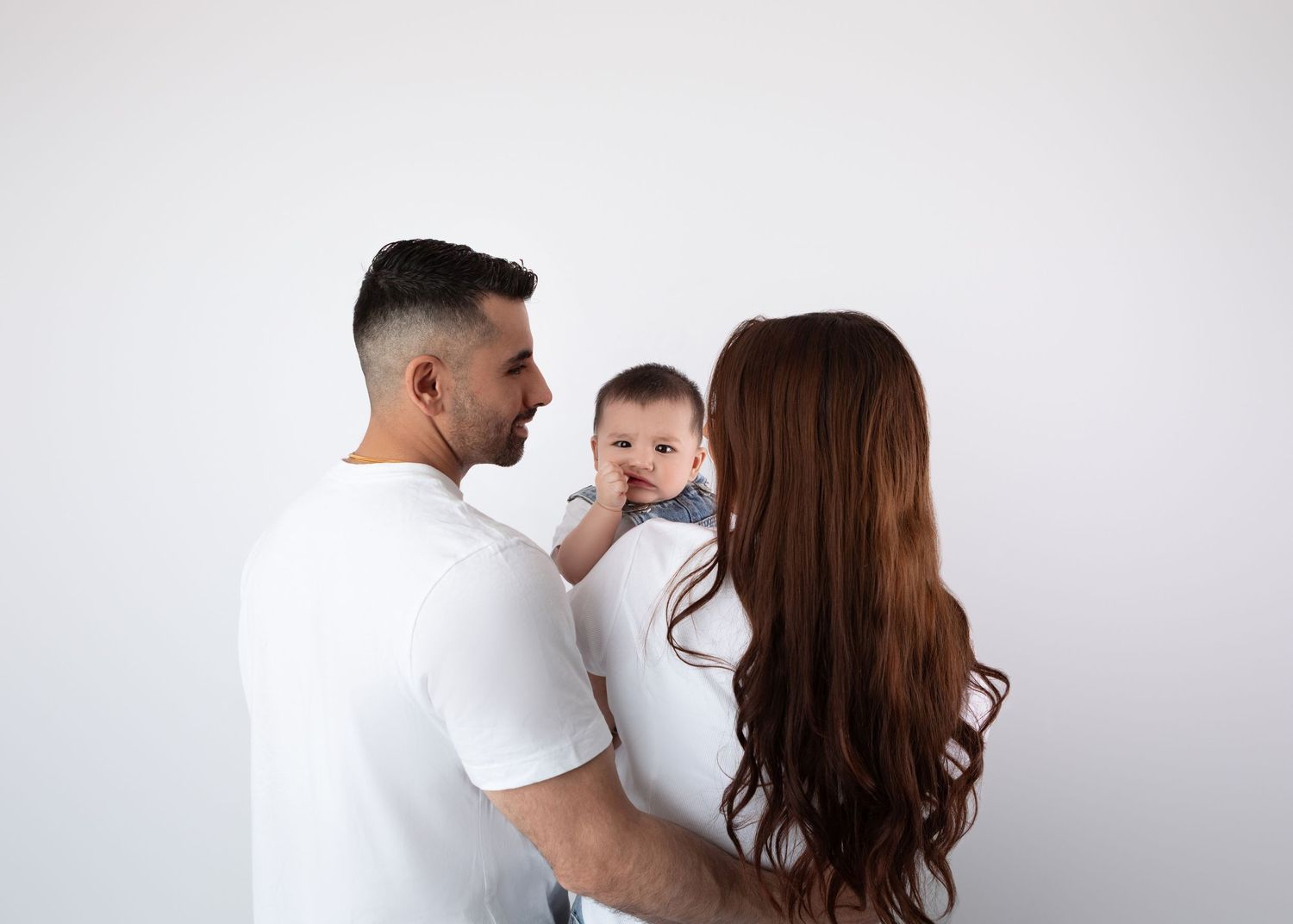 Two parents in white tees lovingly gaze at their baby boy against a white background.