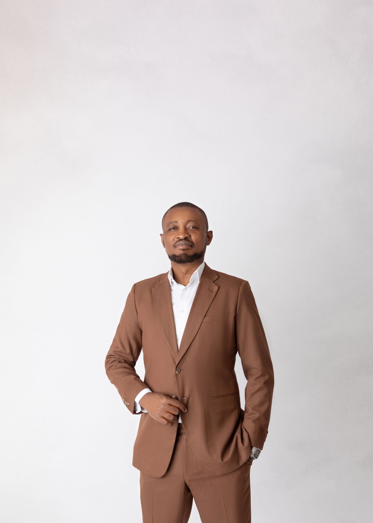 Man in brown suit standing confidently with hands clasped in professional studio portrait.