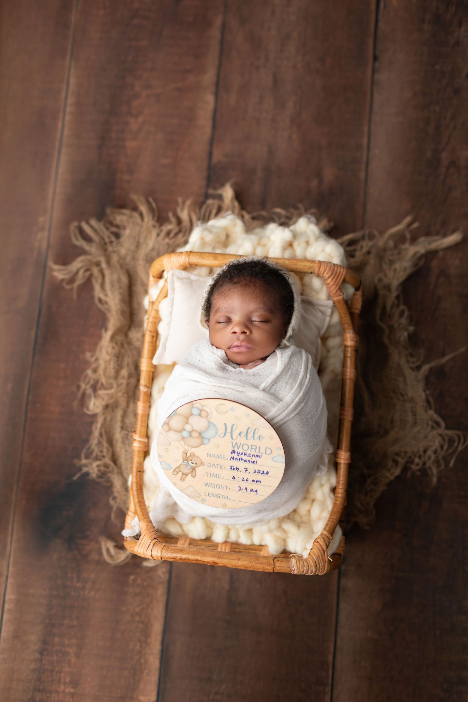 Sleeping newborn in white swaddle lying in wicker bassinet with personalized birth announcement sign.