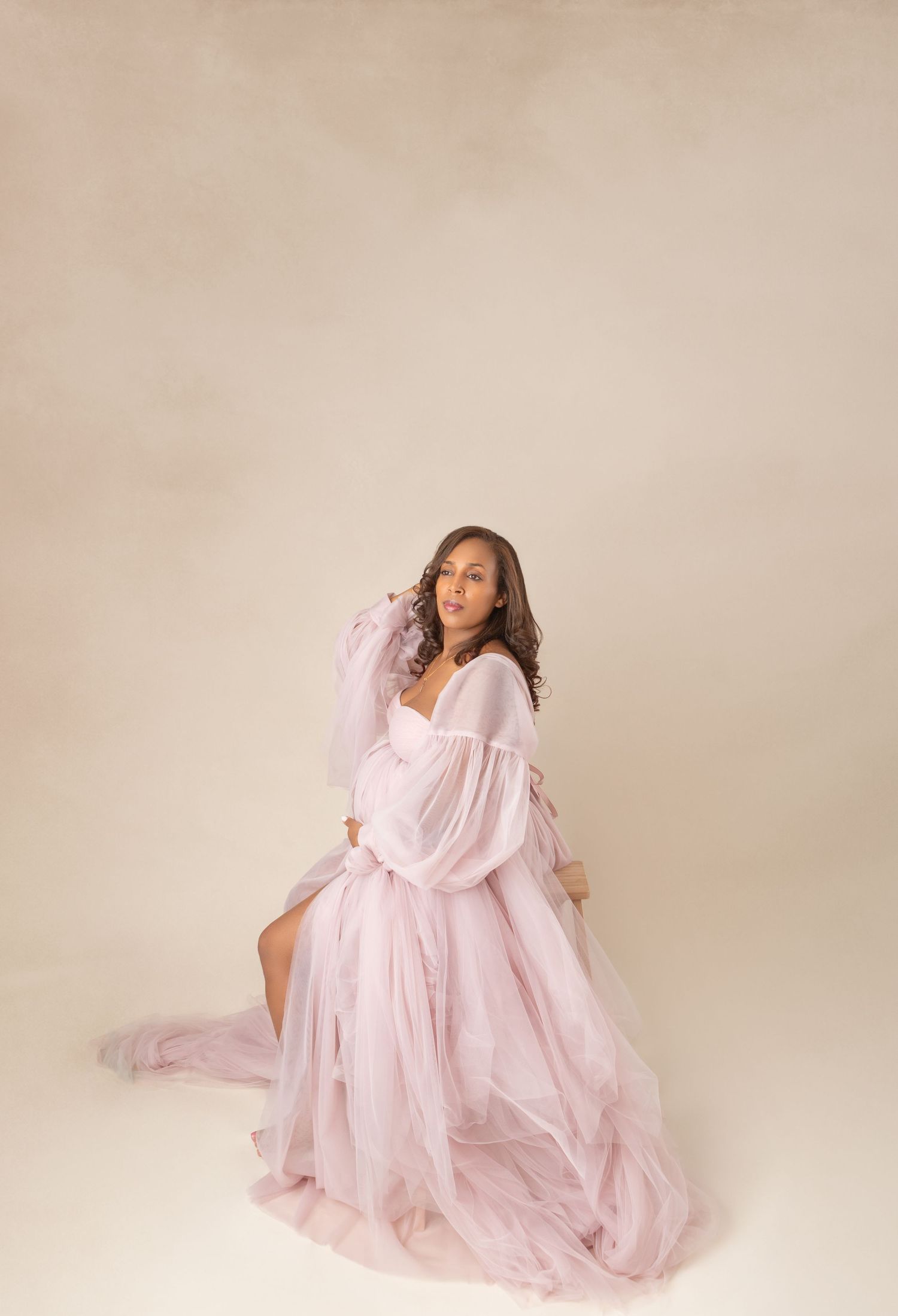 Pregnant woman in blush pink tulle gown sits gracefully in soft-toned studio setting.