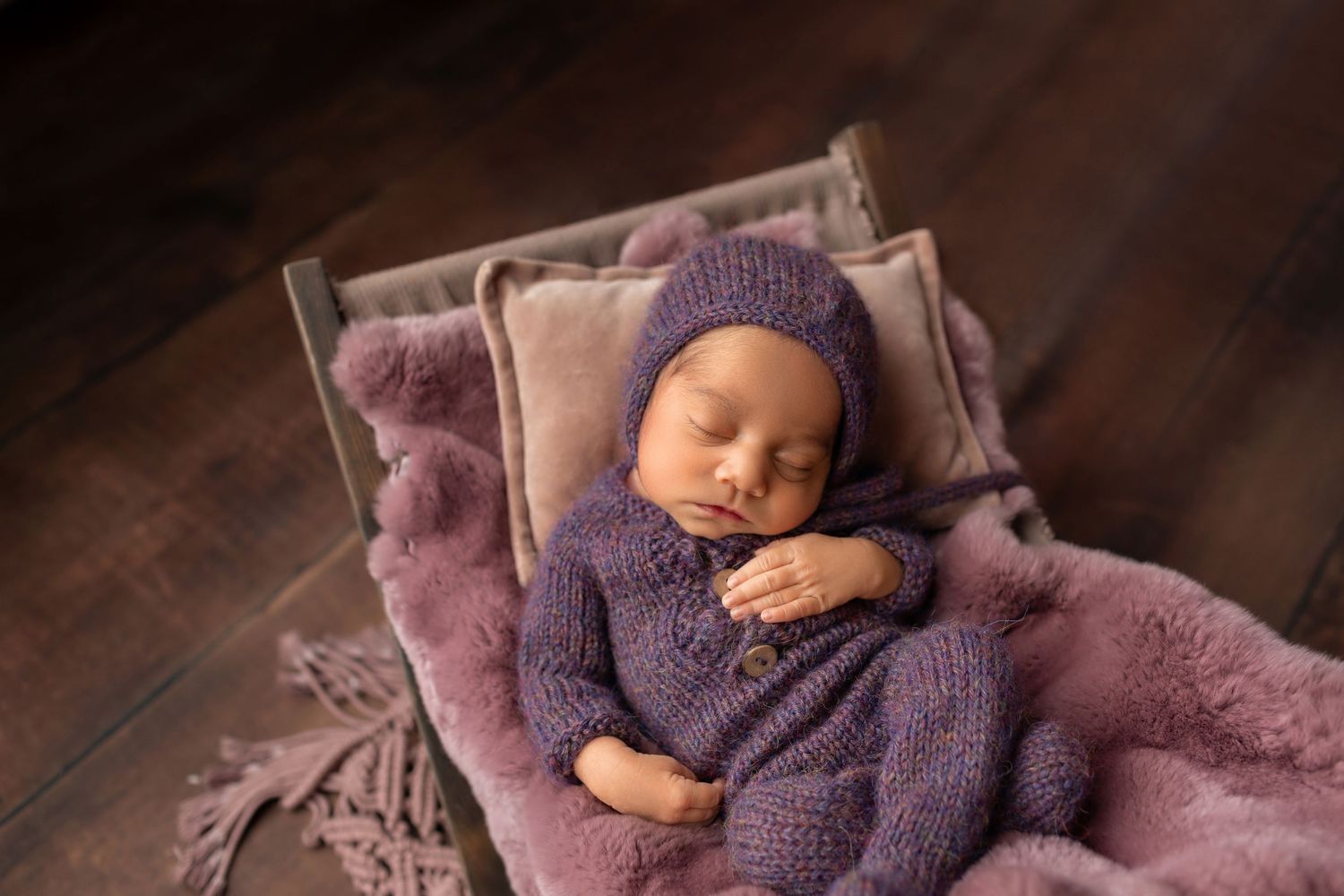 Sleeping newborn baby in purple knit romper and bonnet resting in a tiny wooden bed with mauve blankets.