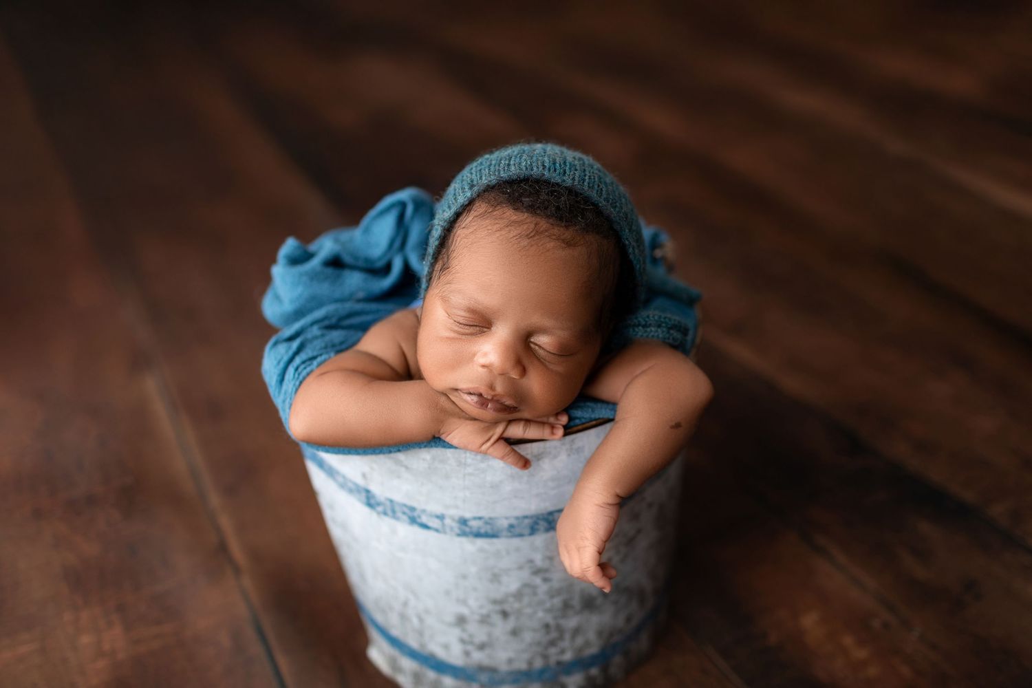 Peaceful newborn baby in blue bonnet resting in small bucket on wood floor.
