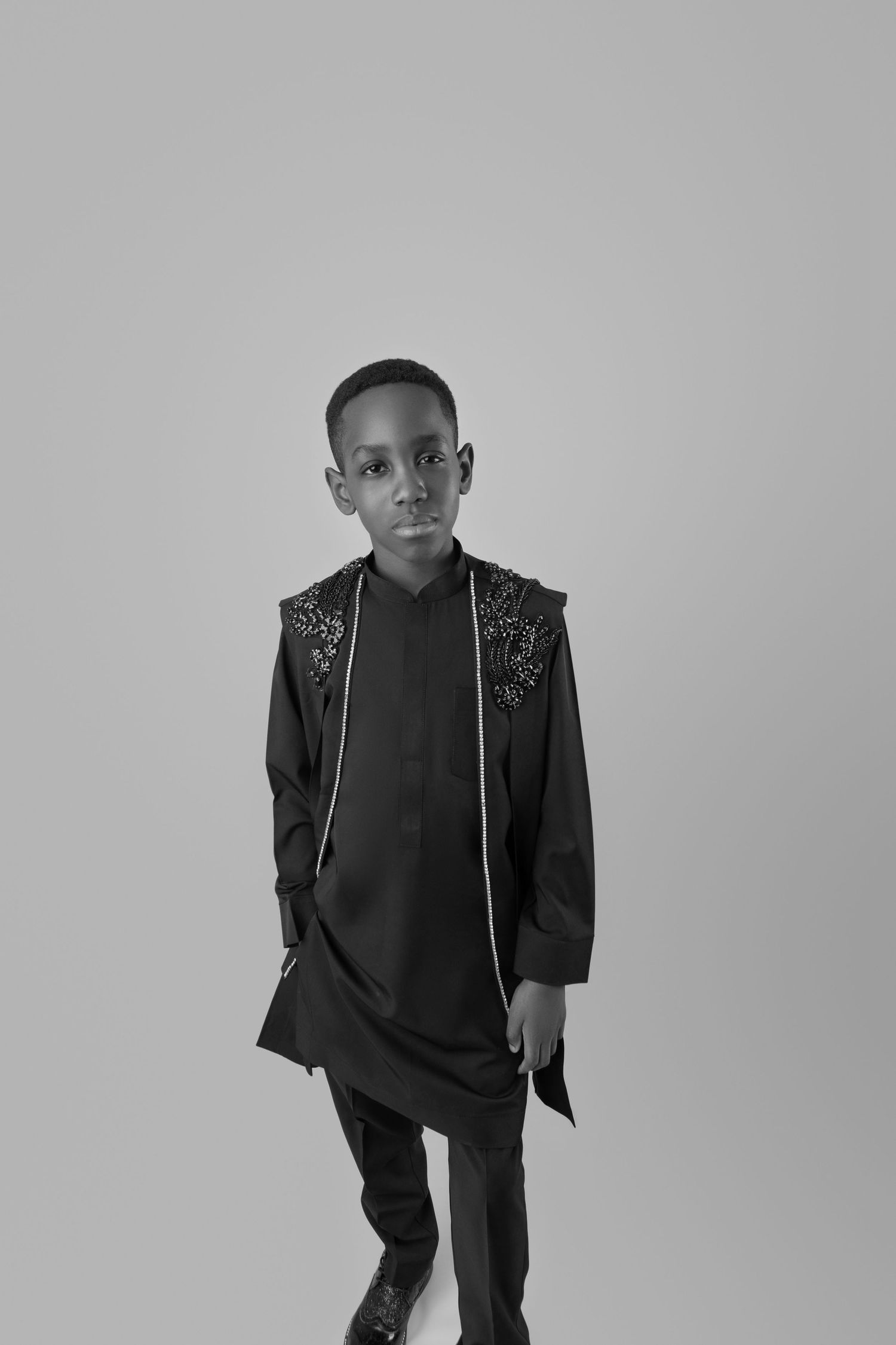 Black and white portrait of young boy wearing ornate black long jacket with embellished shoulders in studio.