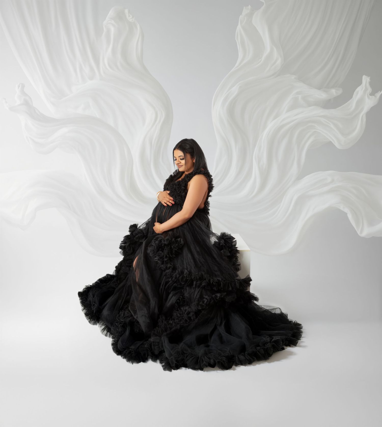 Pregnant woman in elegant black ruffled gown poses gracefully against white swirling backdrop in maternity photo.
