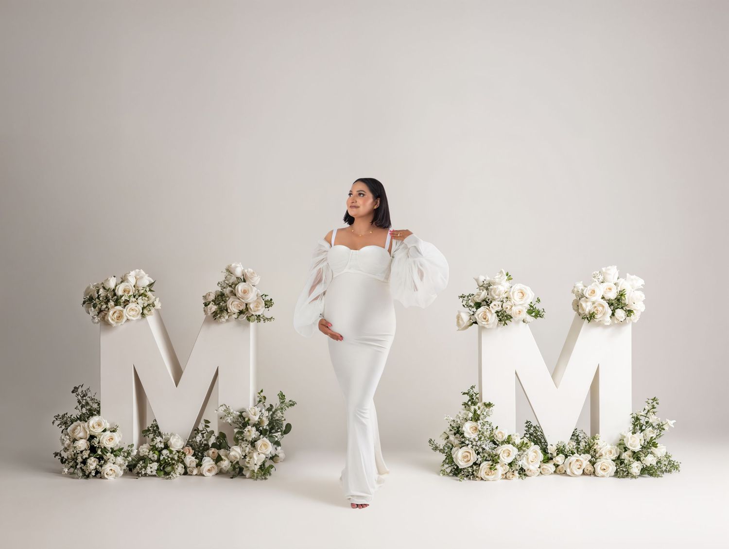 Pregnant woman in white off-shoulder outfit poses between floral M-O-M letters for a maternity photo shoot.