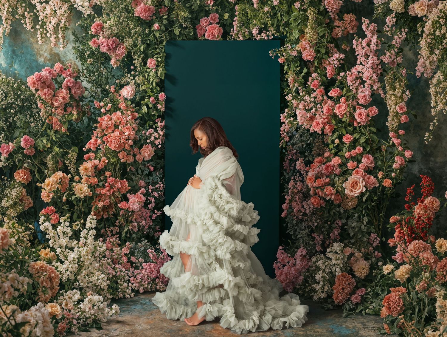 Pregnant woman in white ruffled gown poses gracefully among lush pink flowers against a teal backdrop.