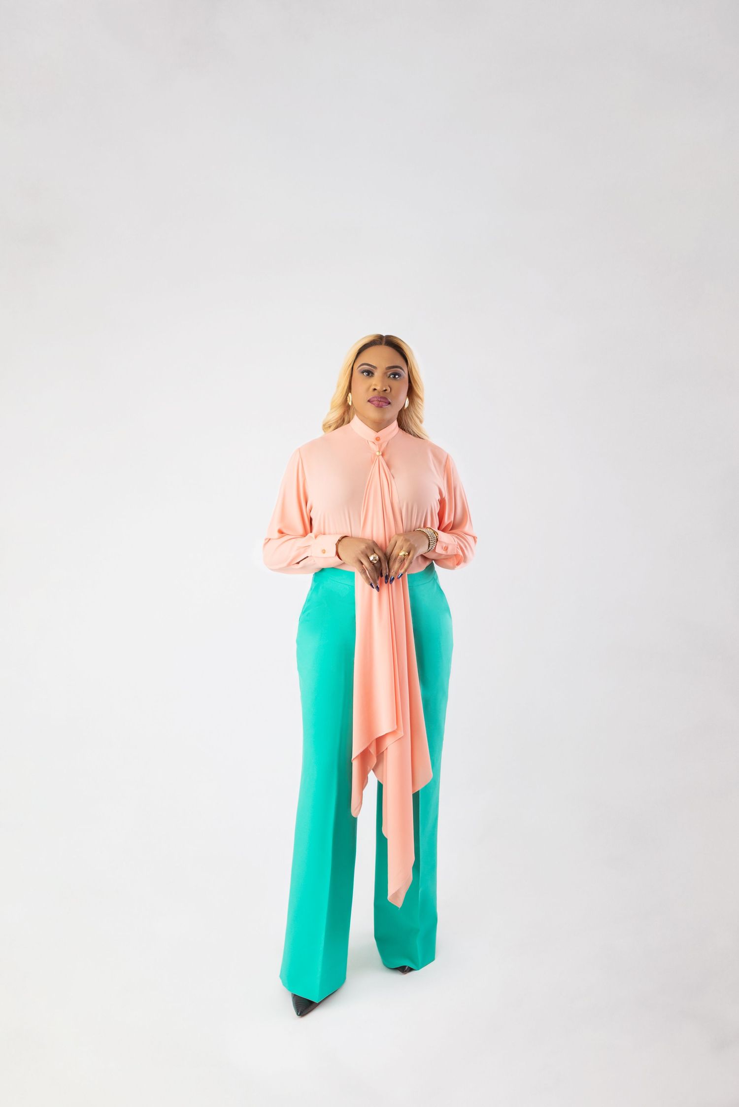 Woman in pink blouse with bow tie and teal wide-leg pants posing against white background.