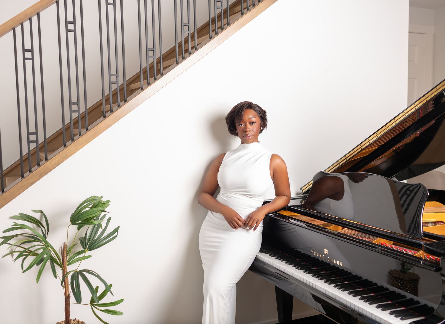 Elegant woman in white gown posing by a grand piano near a staircase with a tropical plant nearby.