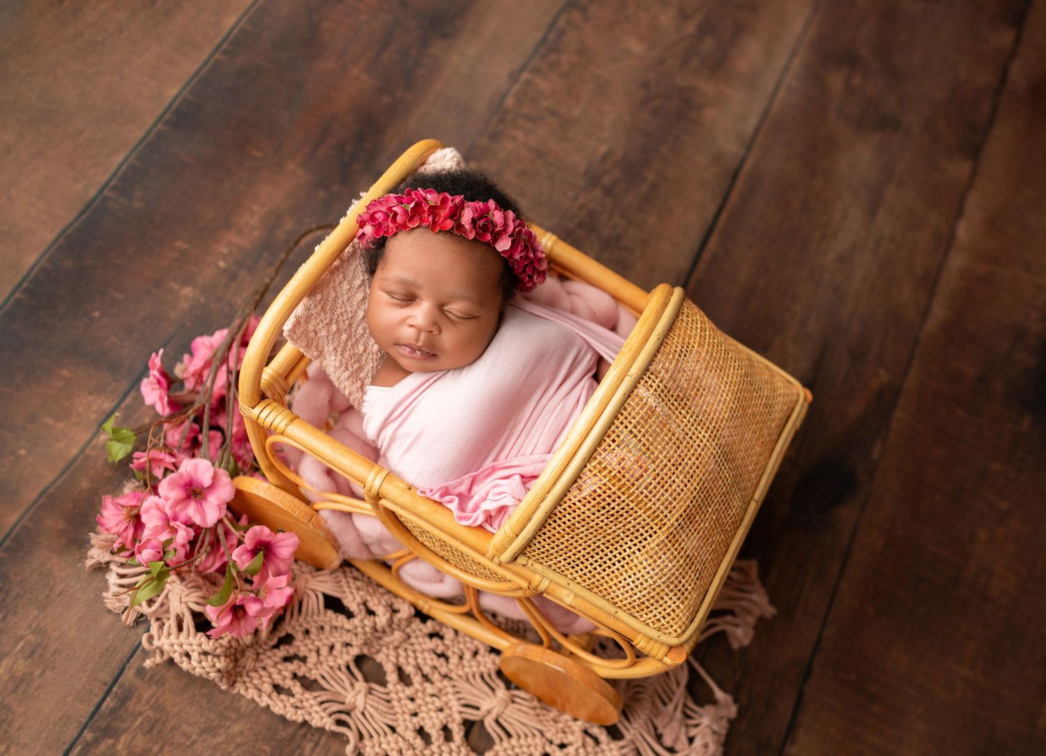 Sleeping newborn baby wrapped in pink swaddle with floral crown resting in wicker basket on wooden floor.