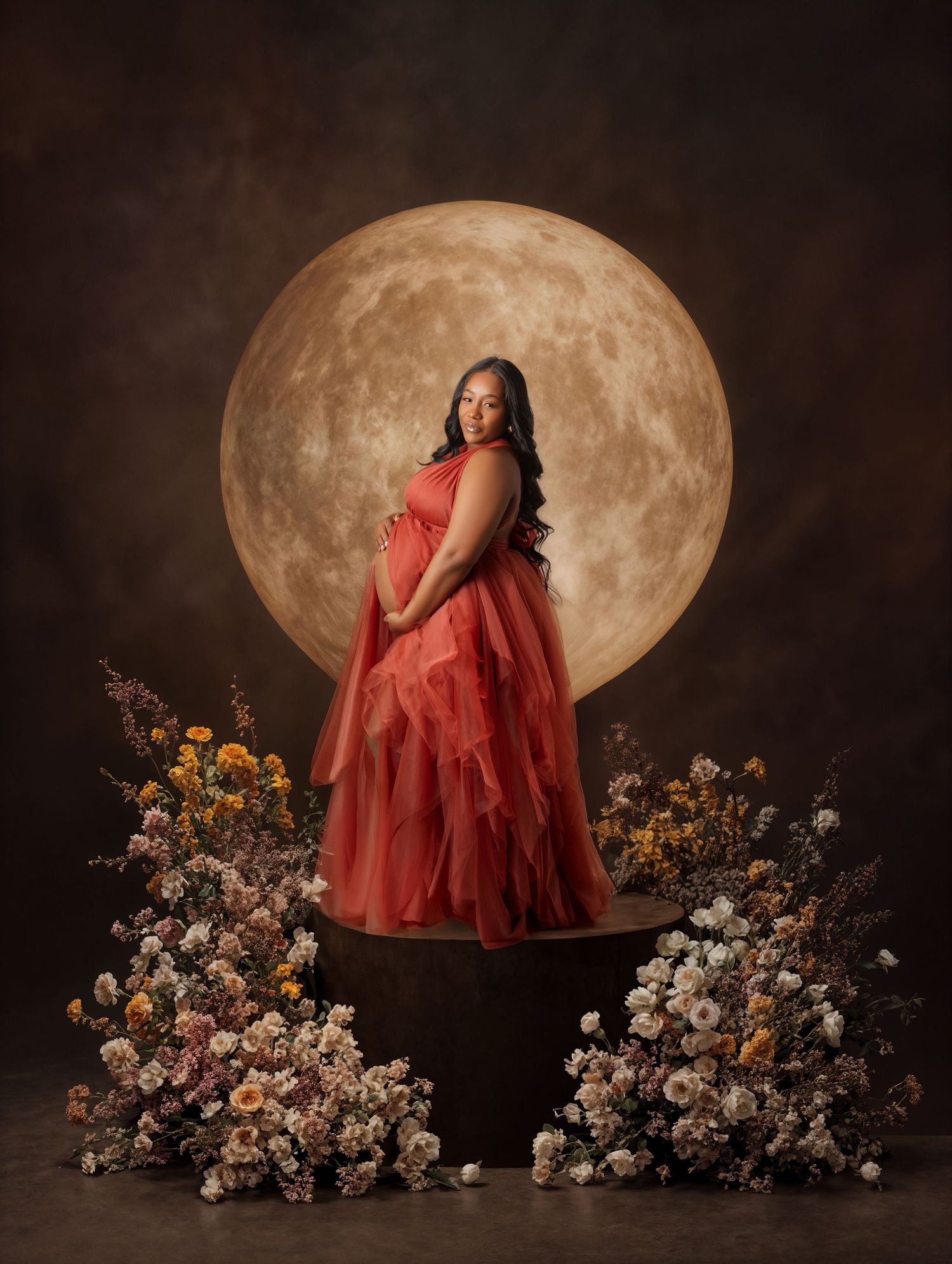 Pregnant woman in red tulle gown stands on pedestal surrounded by flowers before a glowing full moon backdrop.