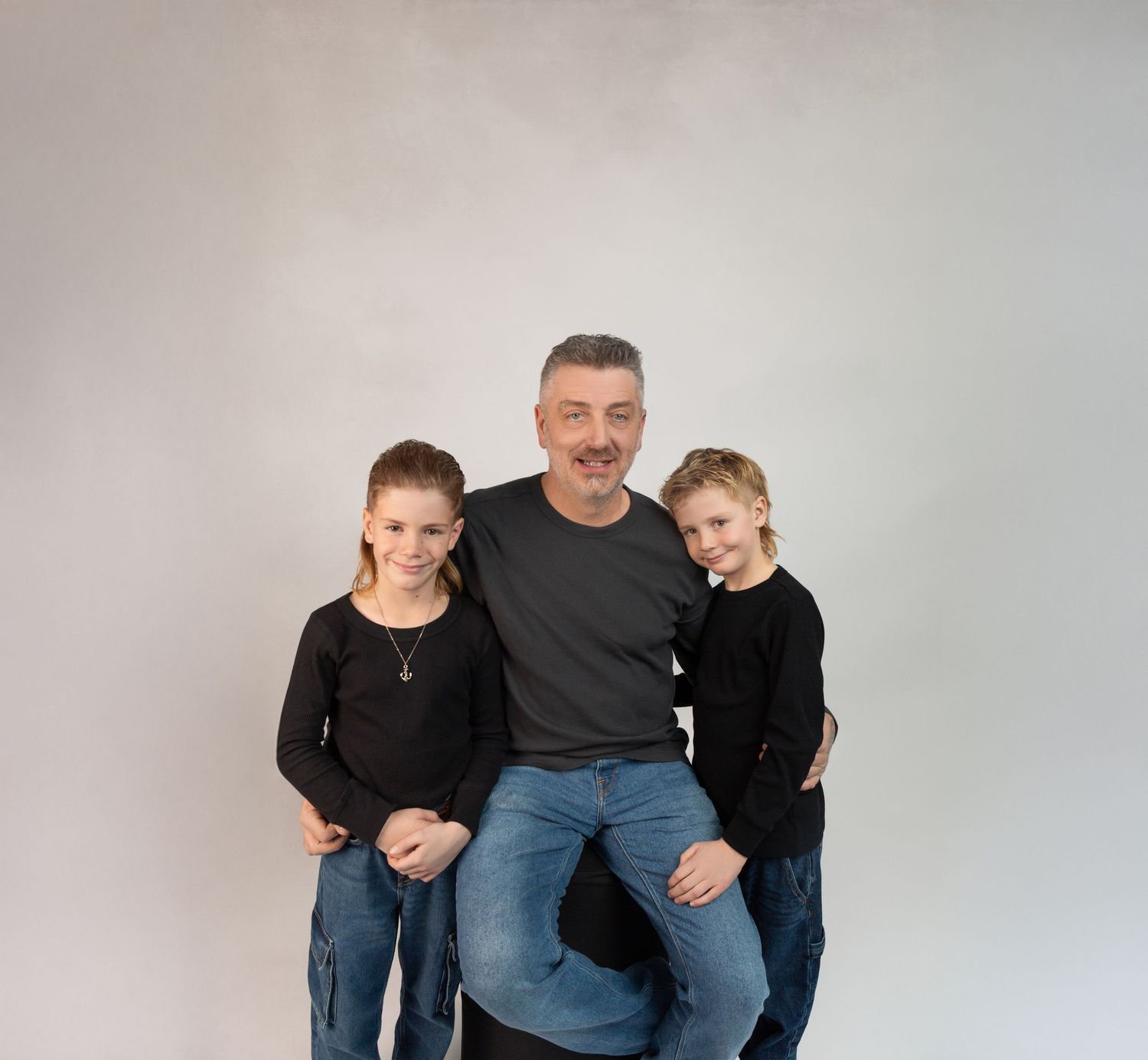 A man in a gray shirt sits with two young girls in black tops on either side of him in a studio portrait.