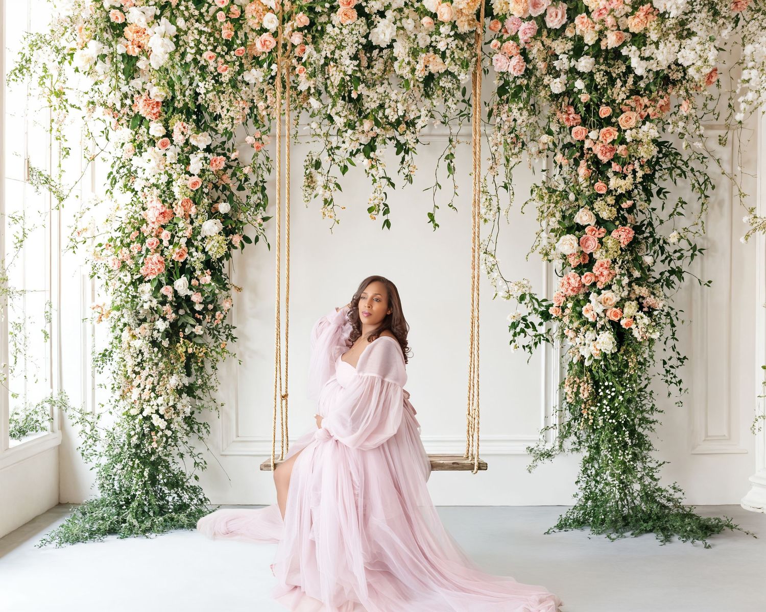 Pregnant woman in pink tulle gown sitting on floral swing surrounded by lush pink and white flower arrangements.