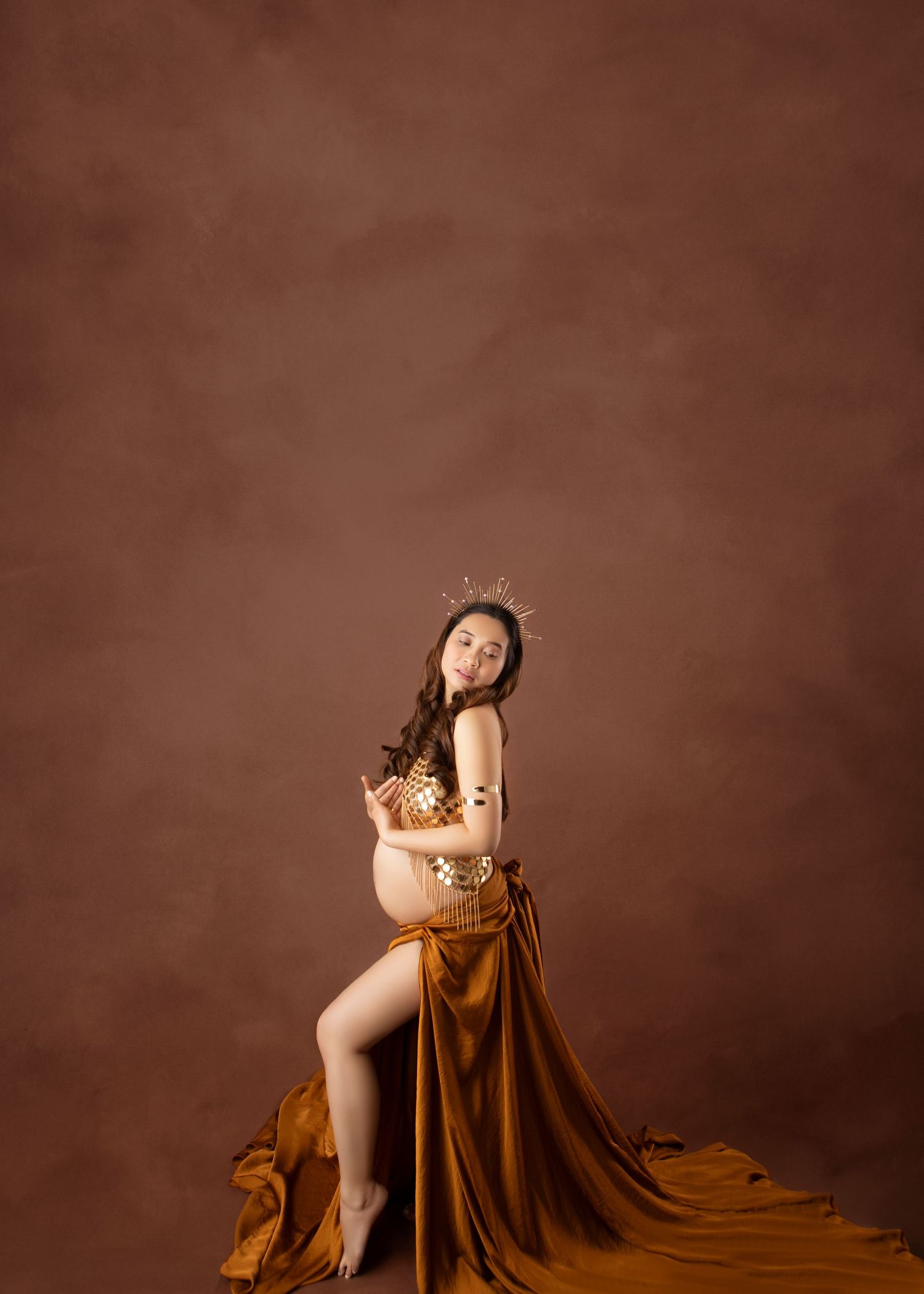 Pregnant woman in gold embellished top and flowing amber skirt with crown posing in studio photography.