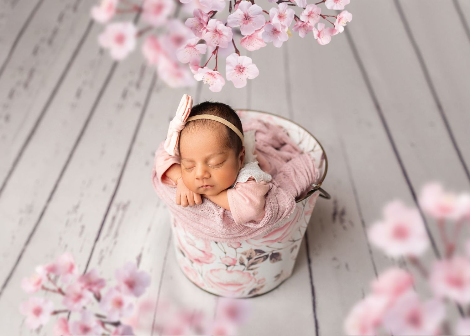 Sleeping newborn in pink wrap inside floral bucket surrounded by cherry blossom branches.