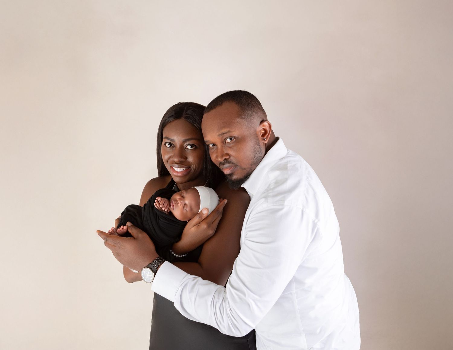 Smiling parents proudly hold their swaddled newborn baby against a warm neutral background.