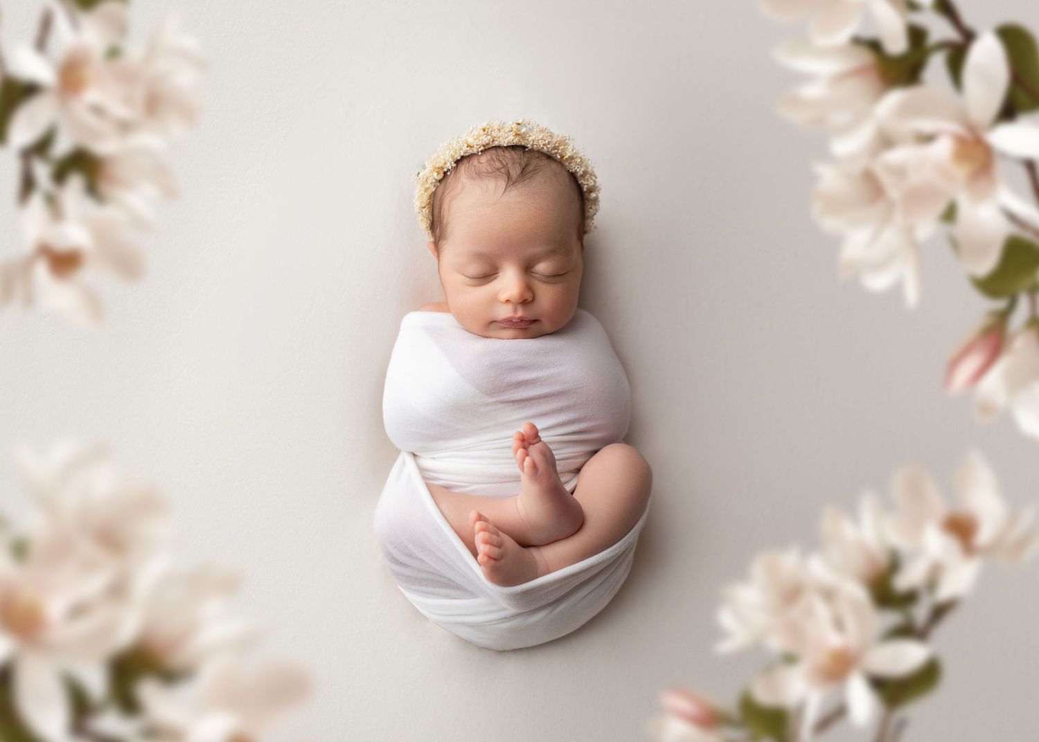 Sleeping newborn wrapped in white swaddle with floral crown, surrounded by soft pink magnolia blooms.