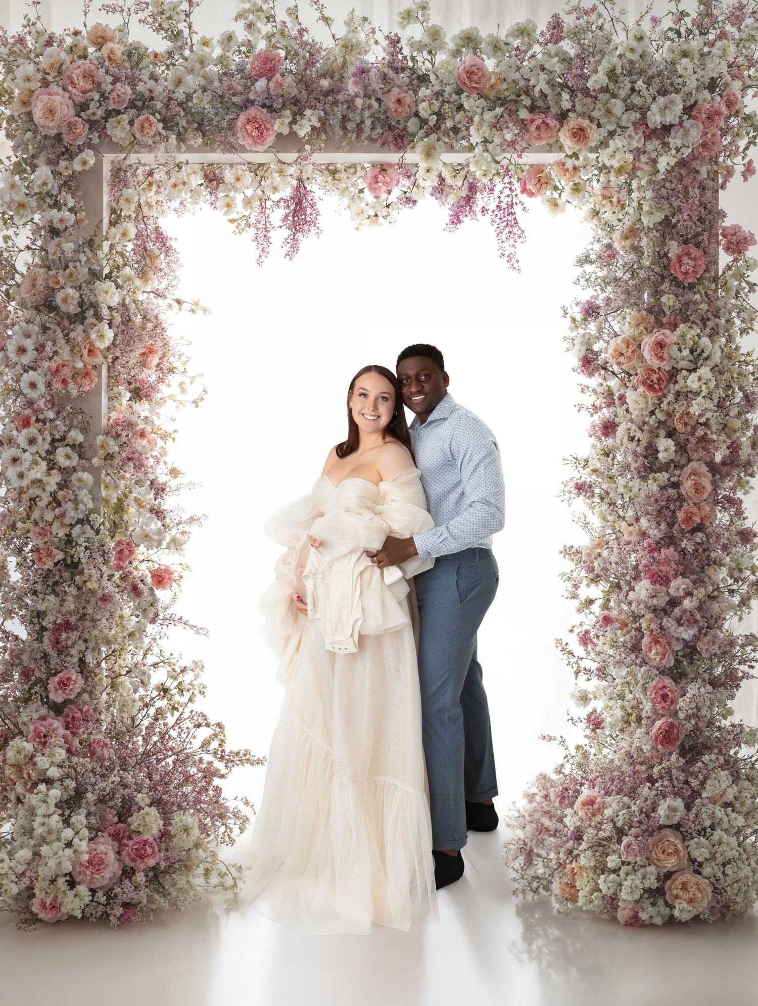 Expecting couple embraces under lush pink floral arch, woman in cream tulle gown, man in blue outfit.