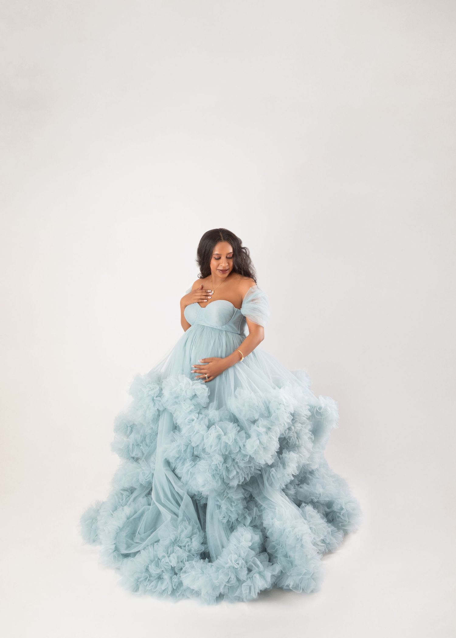 Pregnant woman in a stunning light blue ruffled tulle maternity gown posing gracefully in a white studio.