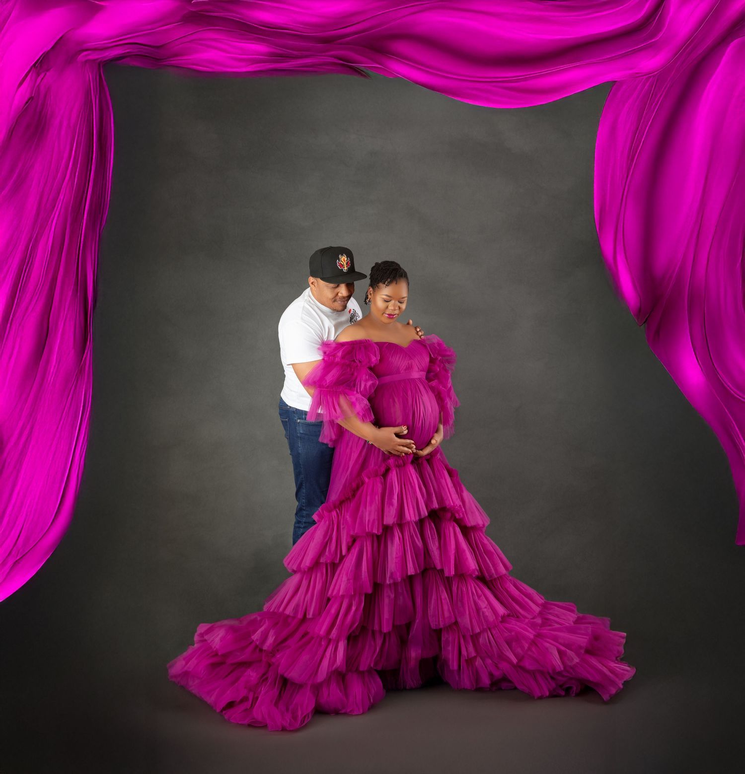 Couple in maternity photo shoot, woman in magenta ruffled gown with pink fabric backdrop on gray background.