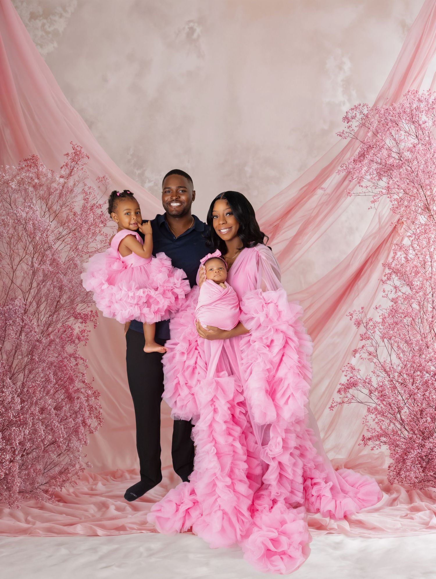Family of four posing in matching pink outfits against a pink floral backdrop in a professional photo shoot.