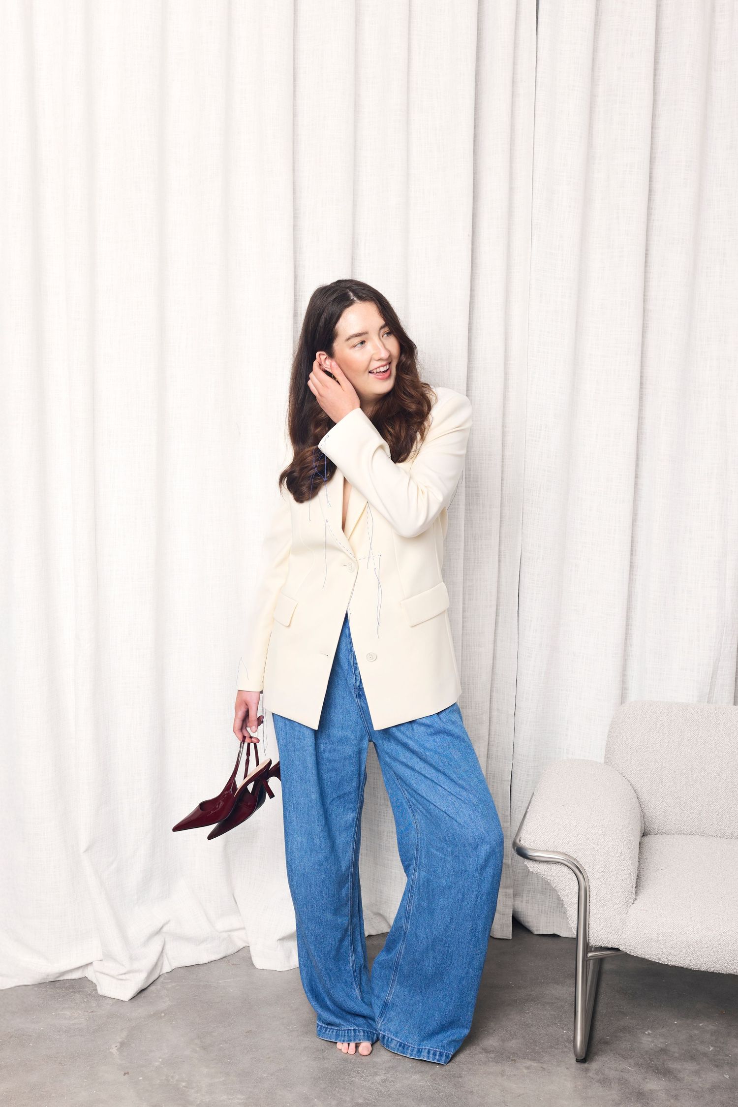 Woman in cream blazer and wide-leg jeans holding heels, posing against white curtain backdrop.