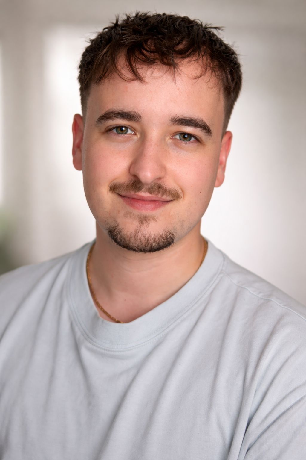 Young man with dark curly hair and beard wearing a light gray t-shirt, smiling in a professional headshot.