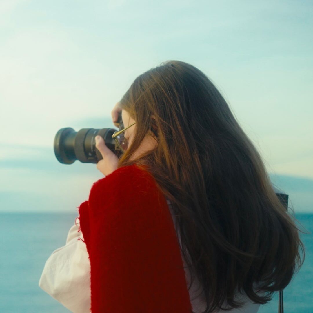 A young woman with long dark hair in a red jacket photographs the ocean with a professional DSLR camera.