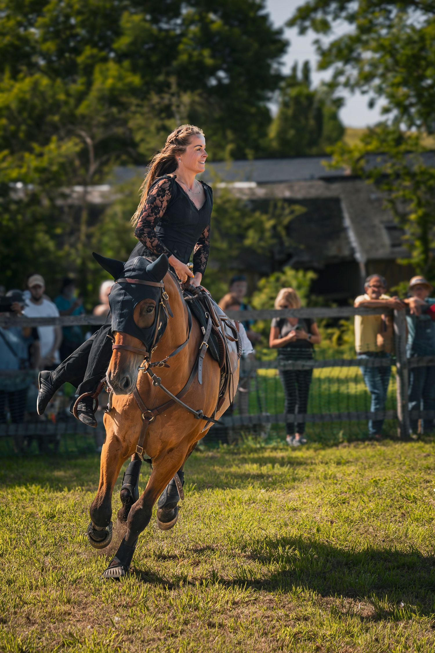Young woman in black dress riding a brown horse rearing up at an outdoor equestrian show with spectators watching.