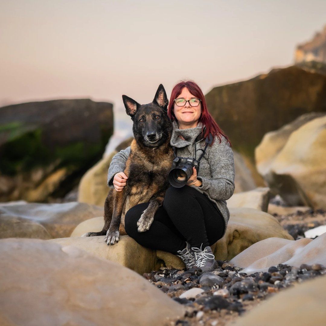 Woman with red hair holds camera while posing with German Shepherd on rocky beach at sunset.
