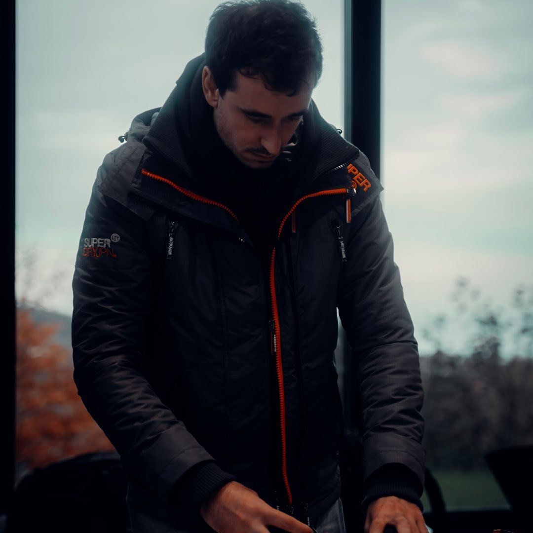 Young man in black and orange Superdry jacket standing near window with autumn trees in background.
