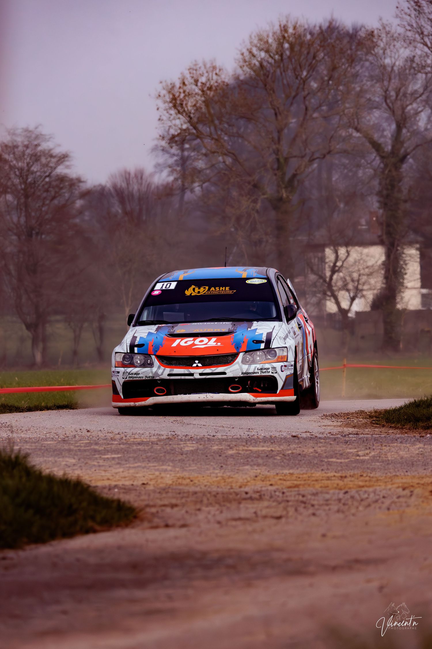 Rally car navigates a dirt corner at high speed during a race through bare winter trees in misty conditions.