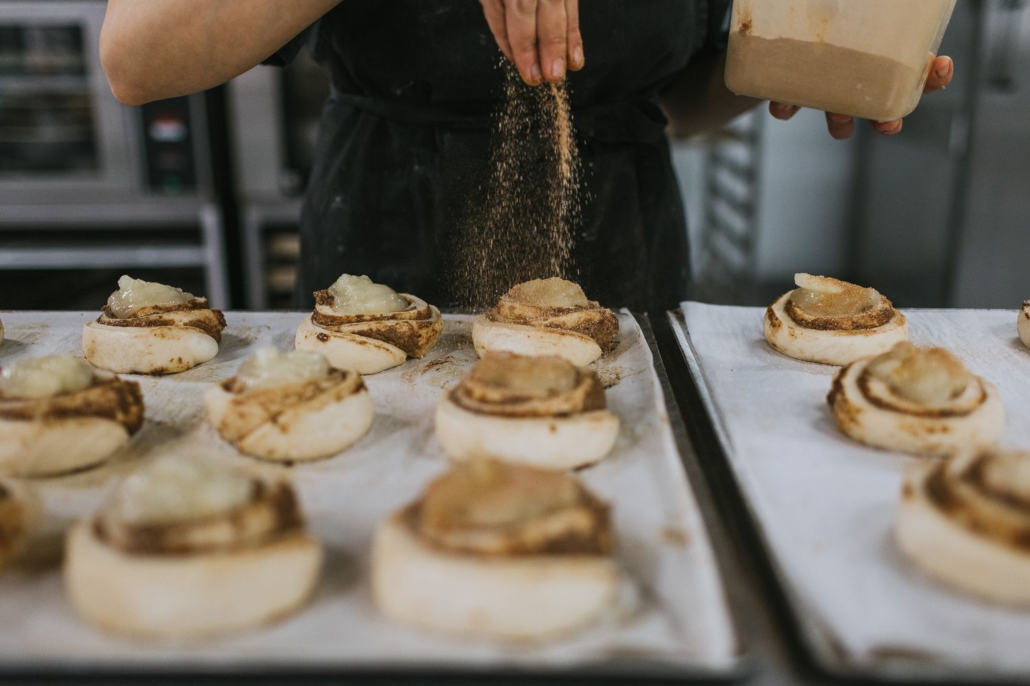 Baker sprinkling sugar on freshly baked cinnamon rolls on a parchment-lined baking tray in a commercial kitchen.