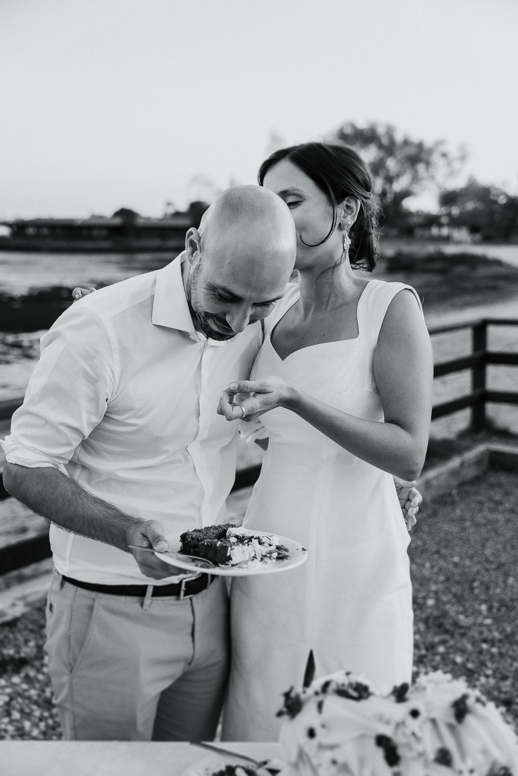 Fotografia de casamento captando um momento doce: a noiva sussurra ao noivo enquanto ele sorri, segurando um prato com bolo.
